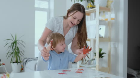 Mom-helps-her-son-to-make-a-handprint-on-paper-using-paint.-Joint-leisure-matter-of-the-child.-Happy-and-caring-mom